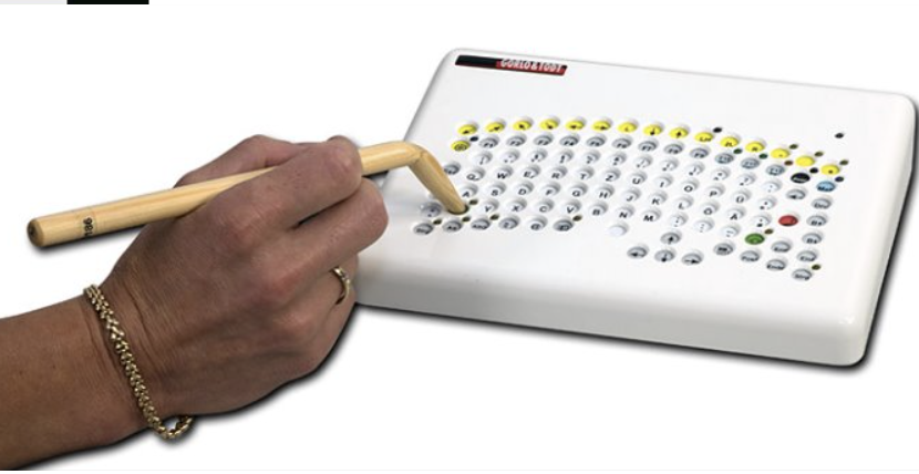 A small keyboard has sunken keys. A hand holds a pointer with an angled end over one of the holes.