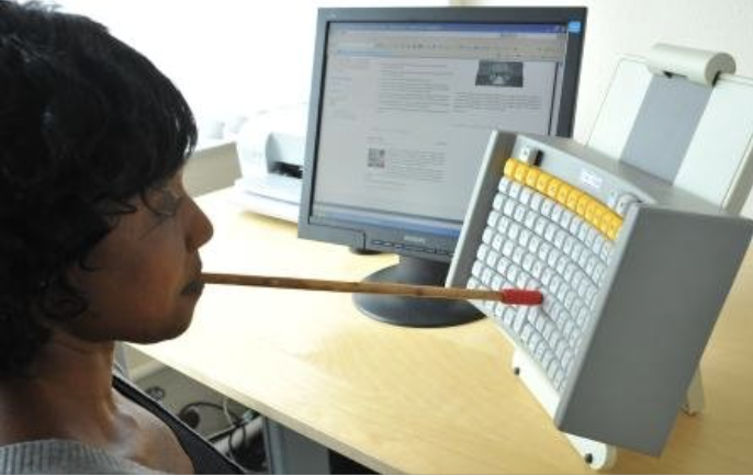 A woman sits at a desk. She uses a stick held in her mouth to press keys on a vertical keyboard in front of her. A screen sits to one side of the keyboard.